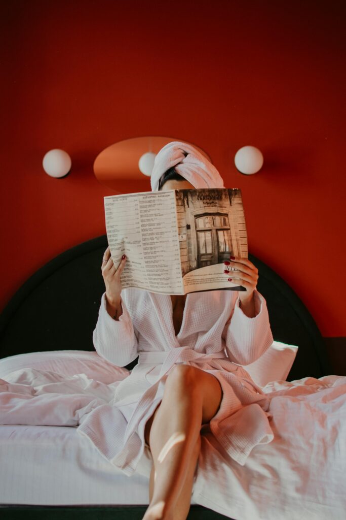 Woman in bathrobe reading a magazine on bed in a cozy hotel room.